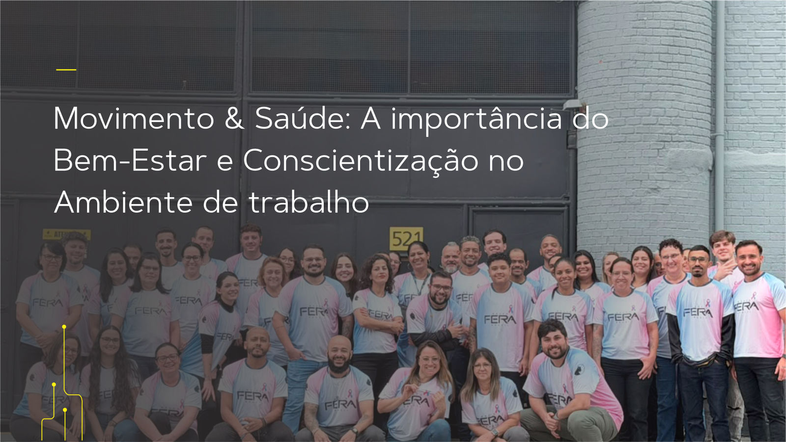 Foto em grupo de colaboradores do Grupo Fera Brasil reunidos ao ar livre, em frente à fachada da empresa com portões industriais cinzas e detalhes em amarelo. Todos vestem camisetas comemorativas nas cores azul e rosa da campanha institucional, sorrindo e olhando para a câmera.